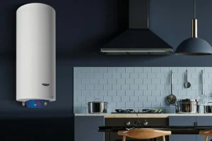 Termo eléctrico Fleck Nilo de 50 litros instalado en la pared de una cocina moderna. El calentador de agua tiene diseño cilíndrico vertical en color blanco, con panel de control digital azul ubicado en la parte inferior. La cocina presenta un estilo contemporáneo con paredes en tonos oscuros, salpicadero de azulejos rectangulares claros y utensilios colgados sobre la encimera. Hay una campana extractora negra y utensilios de cocina de acero inoxidable visibles.