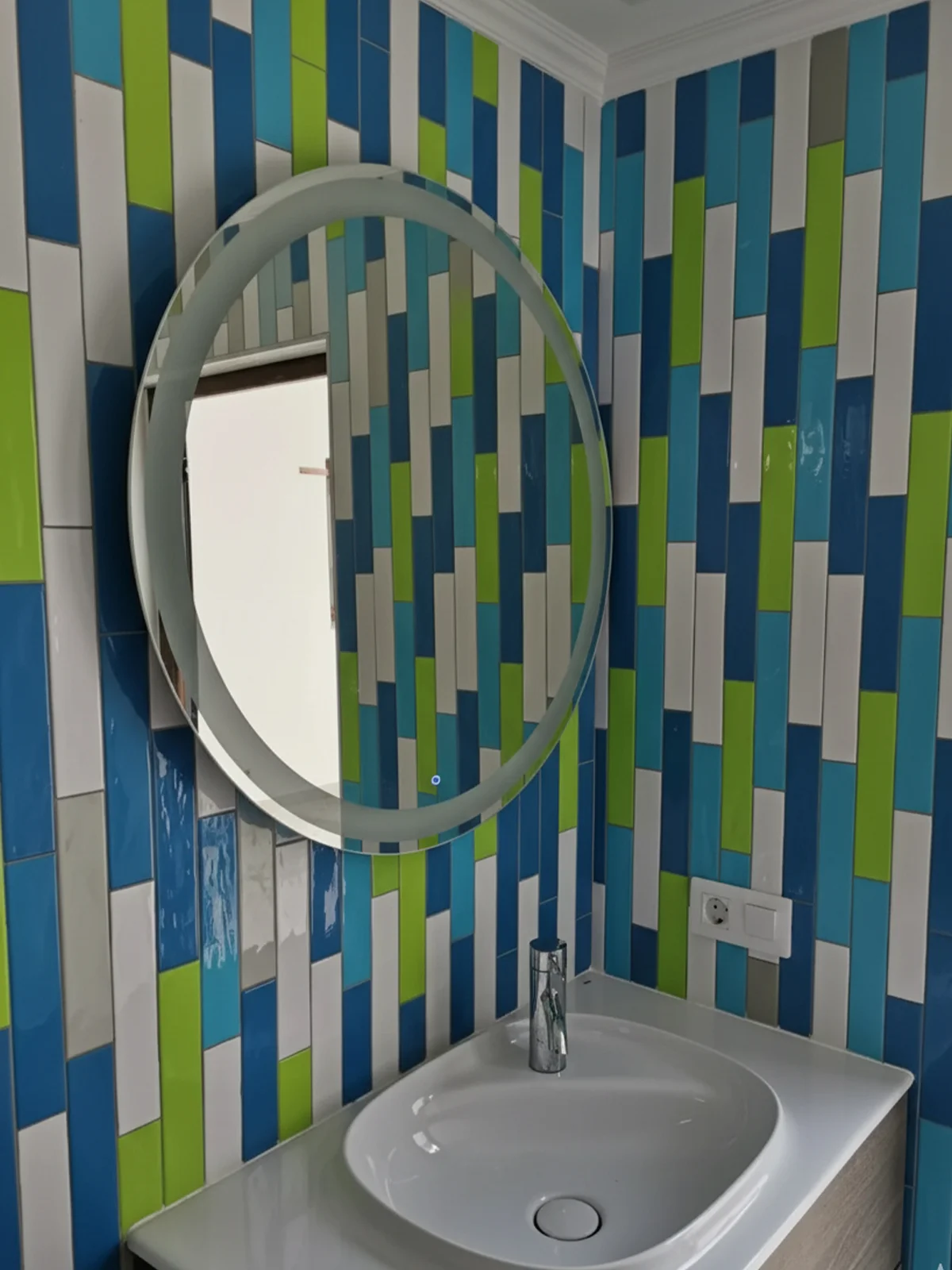 Lavabo moderno con mueble de madera clara, grifo cromado y espejo circular. Paredes decoradas con azulejos verticales en tonos azul, verde, blanco y gris. Reforma de baño en Las Palmas.