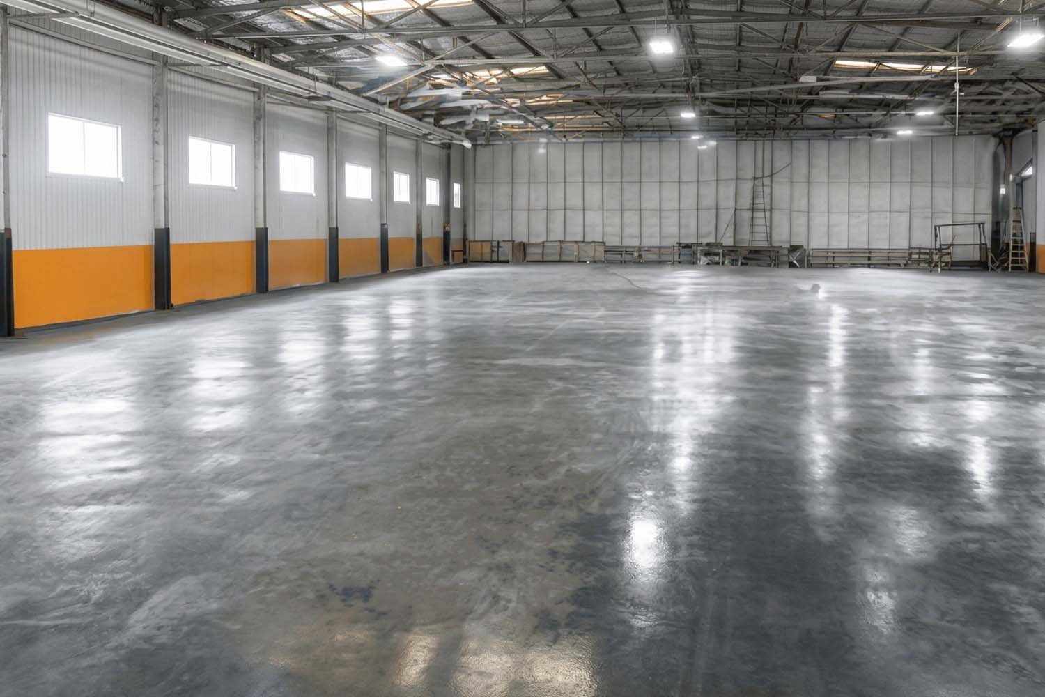 Pavimento en nave industrial con plan de seguridad y salud en obra – Suelo de hormigón pulido industrial En la imagen se observa el pavimento continuo de hormigón pulido en una nave industrial de gran tamaño, con acabado brillante y superficie nivelada, diseñado para uso logístico o industrial. El espacio interior presenta estructura metálica vista, iluminación superior y amplias paredes laterales, cumpliendo criterios de plan de seguridad y salud en obra para pavimento en nave industrial.